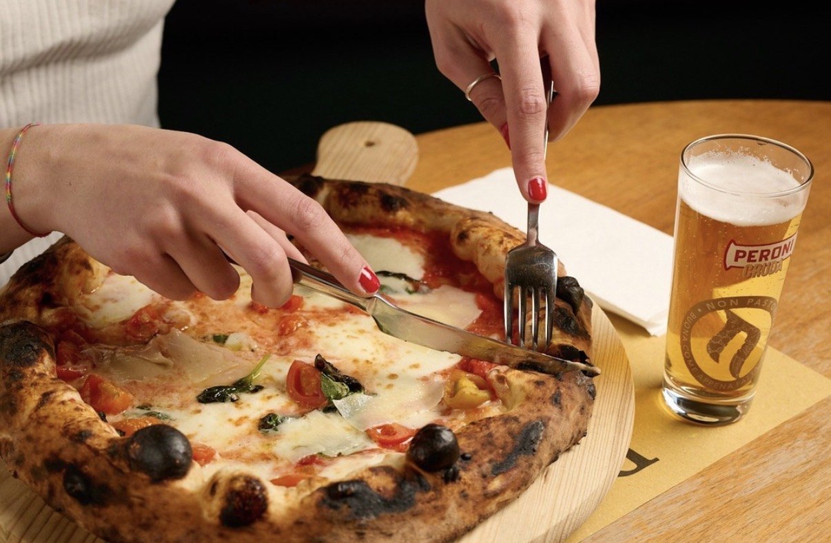 Mani che tagliano una pizza margherita su tagliere di legno accanto a una birra Peroni
