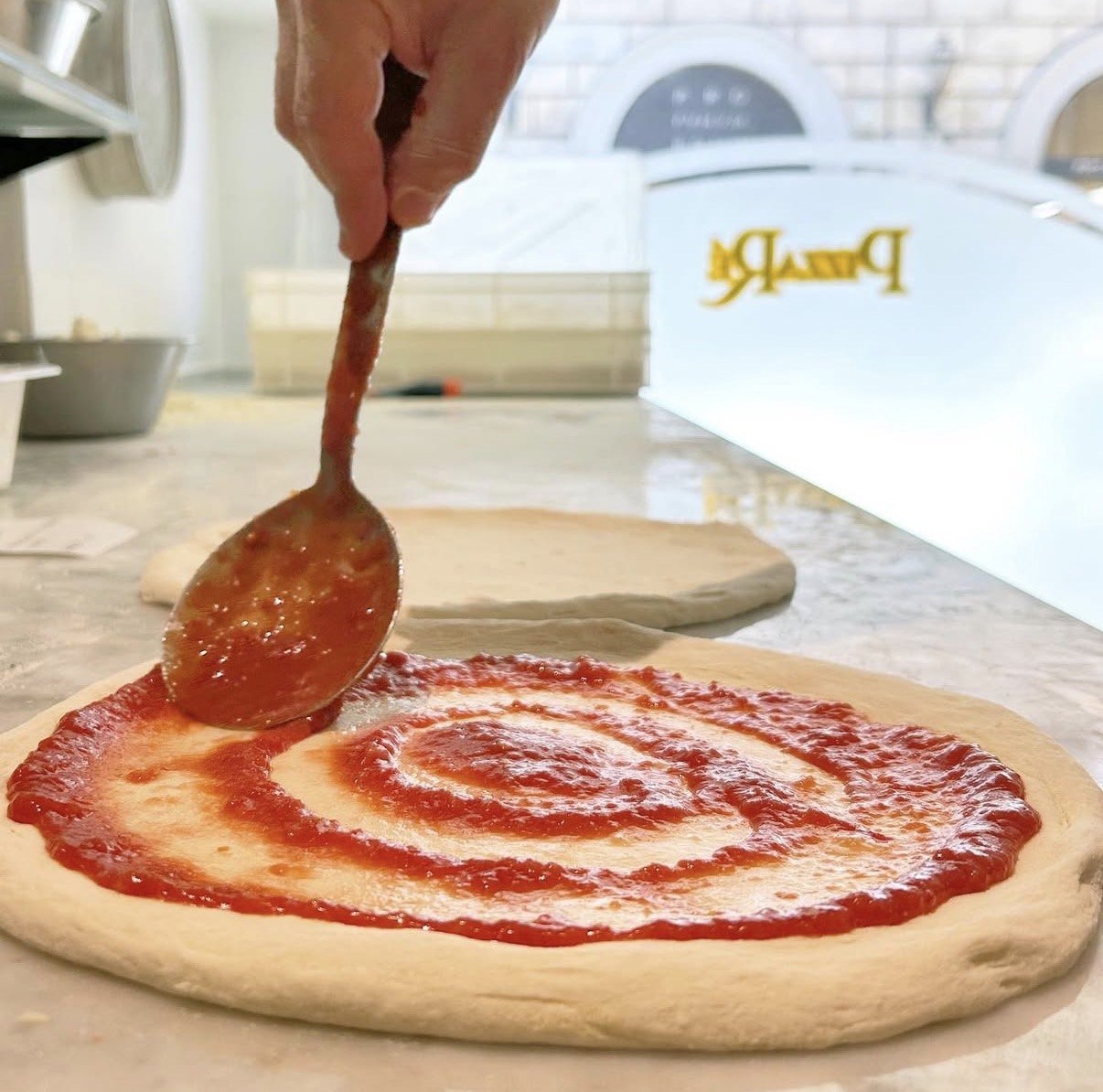 Pizzaiolo PizzaRé che stende a cucchiaio la salsa di pomodoro sul disco di impasto davanti al forno